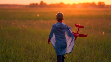 The-boy-in-the-costume-of-a-super-hero-running-in-a-red-cloak-laughing-at-sunset-in-summer-field-representing-that-he-was-the-pilot-of-the-plane-playing-with-a-model-airplane